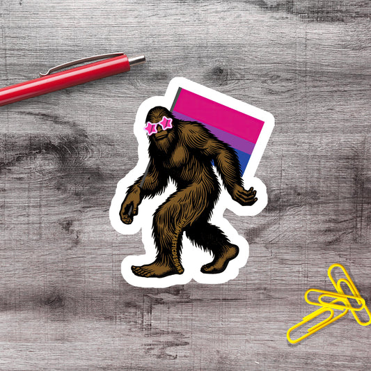 a sticker of a cartoon Sasquatch wearing sunglasses and carrying a flag, along with a red pen and yellow paper clips on a wooden surface.