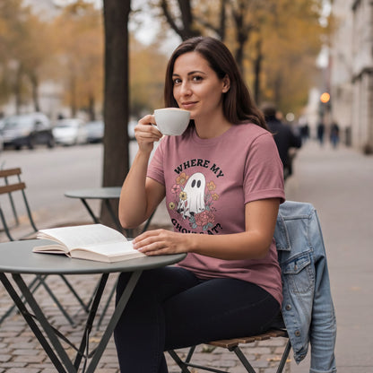 A woman wearing a pink t-shirt with a ghost graphic is sitting at a table, reading a book and drinking from a cup.