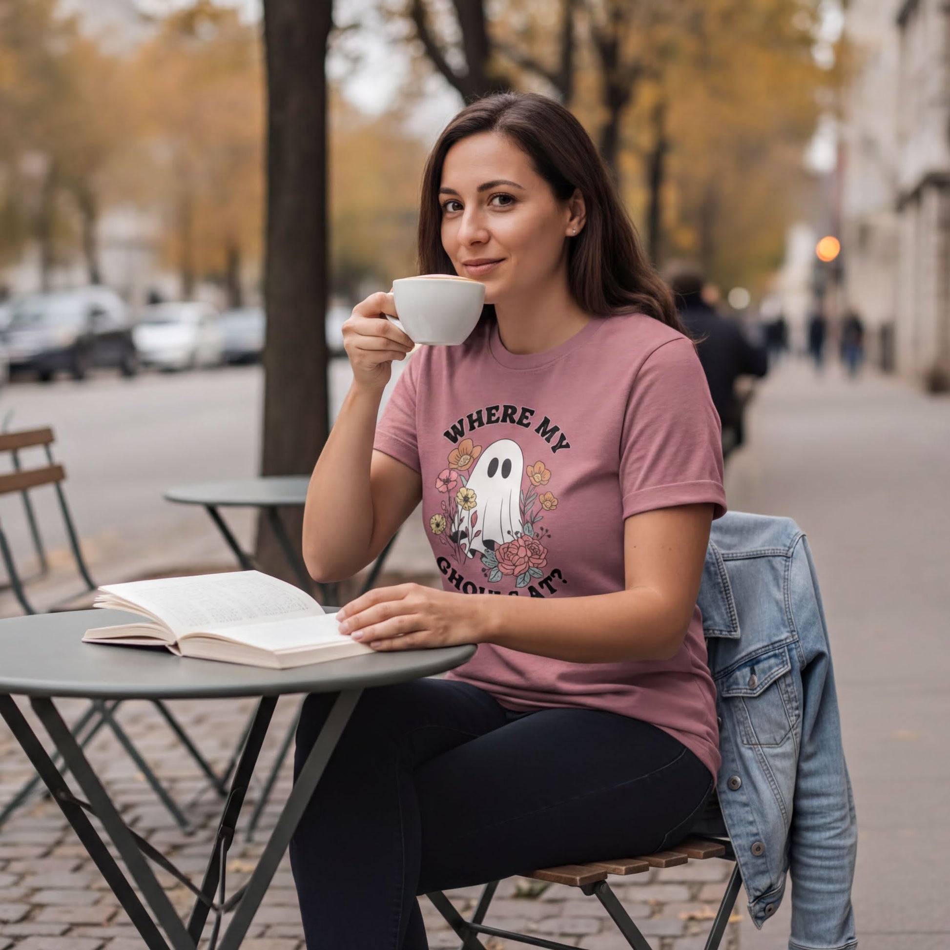 A woman wearing a pink t-shirt with a ghost graphic is sitting at a table, reading a book and drinking from a cup.