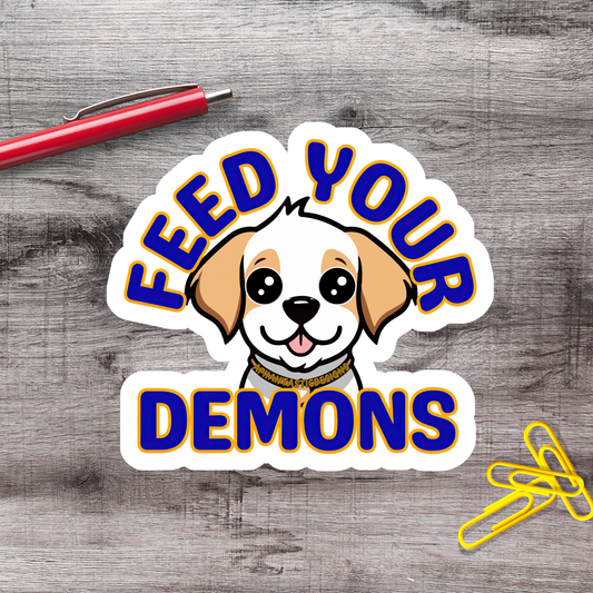 a sticker with a cartoon dog and the text "FEED YOUR DEMONS" on a wooden surface.