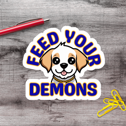 a sticker with a cartoon dog and the text "FEED YOUR DEMONS" on a wooden surface.