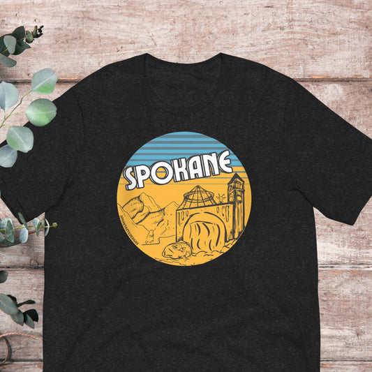 A black t-shirt with a graphic design featuring a circular illustration of a city skyline, including a bridge and mountains, with the word "SPOKANE" prominently displayed.