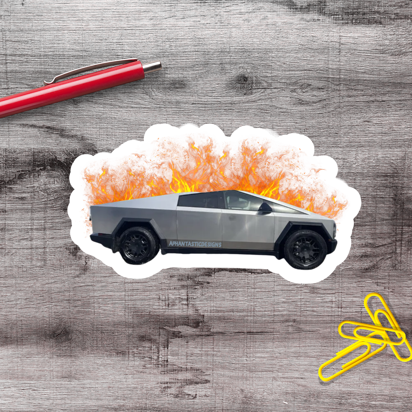 A sticker depicting a silver truck with flames coming out of the hood, placed on a wooden surface alongside a red pen and yellow paper clips.