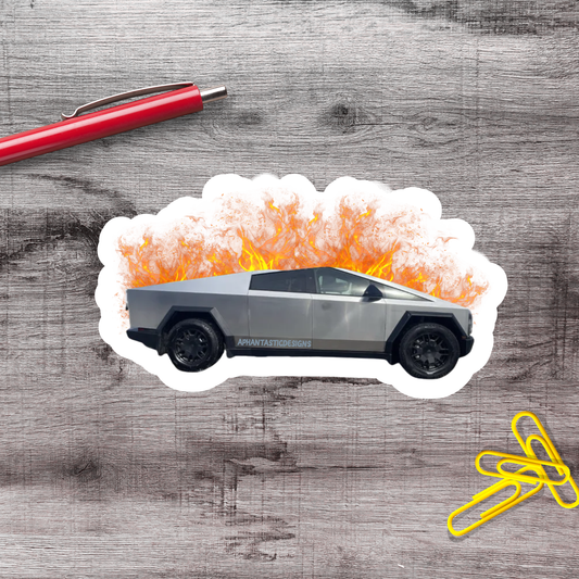 A sticker depicting a silver truck with flames coming out of the hood, placed on a wooden surface alongside a red pen and yellow paper clips.