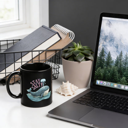 a desk with a laptop, a black coffee mug with a whale design, a notebook, a potted plant, and a seashell.