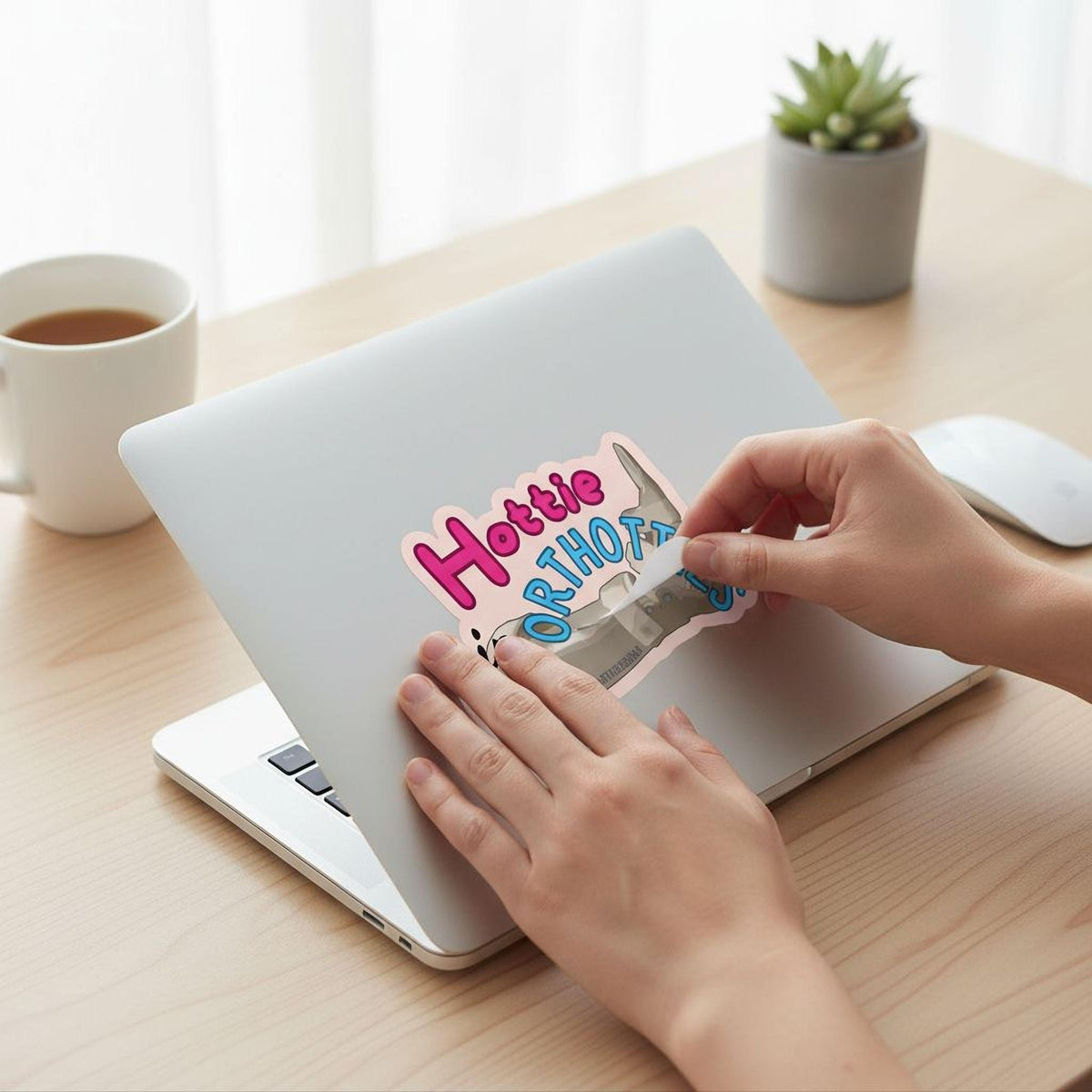 A person's hands are placing a sticker on a laptop computer, which is placed on a wooden desk.