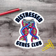 a sticker with the text "DISTRESSAED GENES CLUB" and an illustration of a pair of jeans with a DNA helix. There is also a red pen and a pile of yellow paper clips on a wooden surface.