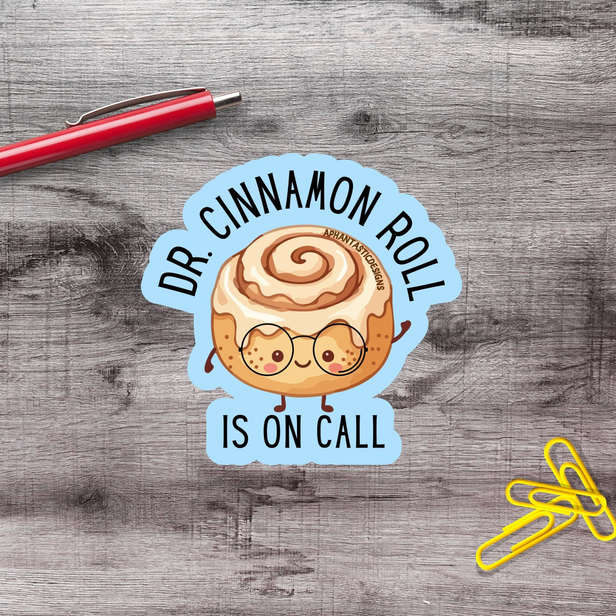 A sticker with a cinnamon roll character and the text "Dr. Cinnamon Roll is on Call" is placed on a wooden surface next to a red pen and yellow paper clips.