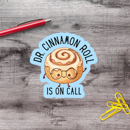 A sticker with a cinnamon roll character and the text "Dr. Cinnamon Roll is on Call" is placed on a wooden surface next to a red pen and yellow paper clips.
