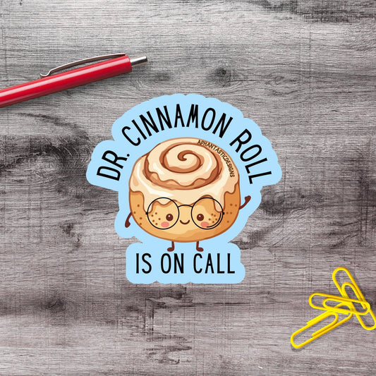 A sticker with a cinnamon roll character and the text "Dr. Cinnamon Roll is on Call" is placed on a wooden surface next to a red pen and yellow paper clips.