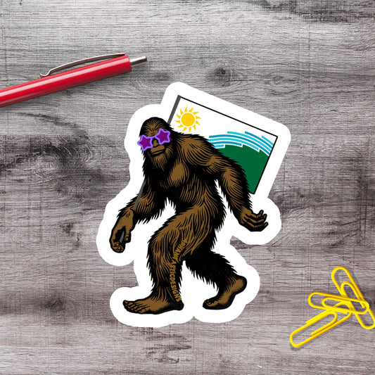 A sticker of a cartoon Sasquatch wearing sunglasses and standing on a piece of paper with a landscape and sun on it, along with a red pen and yellow paper clips on a wooden surface.