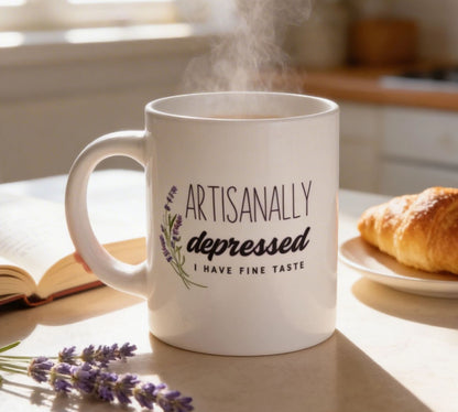 A white mug with the text "Artisanally Depressed I HAVE FINE TASTE" printed on it, placed on a table next to a plate of food and a book.