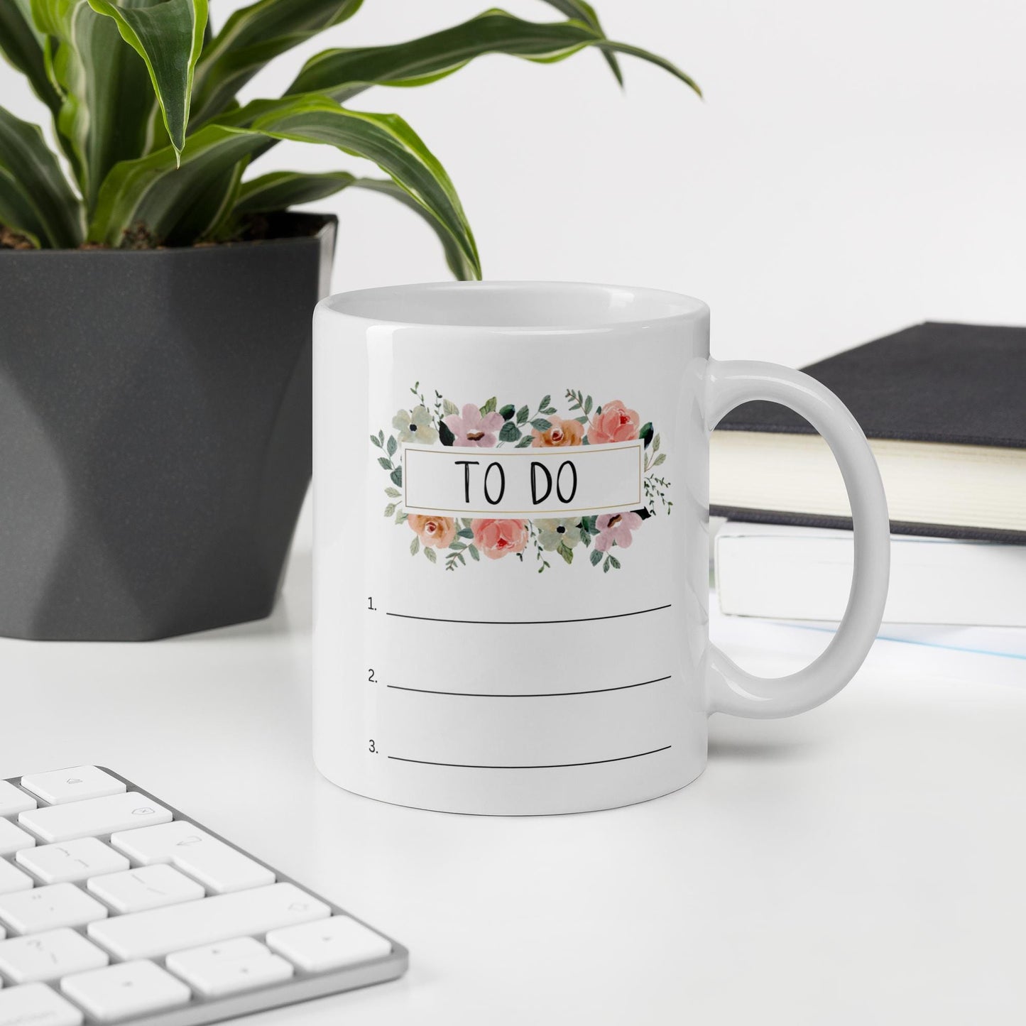 A white mug with a floral design and the text "TO DO" sits on a desk, with a keyboard and a plant in the background.