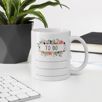 A white mug with a floral design and the text "TO DO" sits on a desk, with a keyboard and a plant in the background.