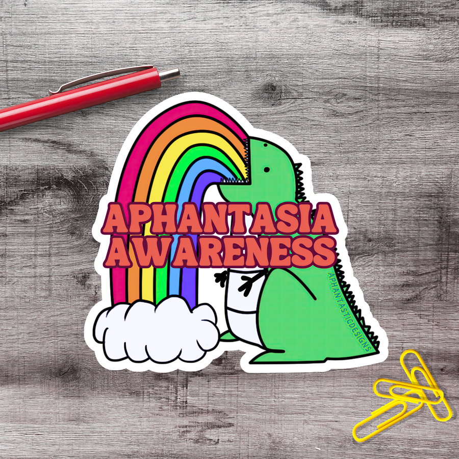 A sticker with a green dinosaur and a rainbow, along with the text "Aphantasia Awareness" on a wooden surface.
