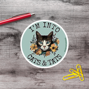 A sticker featuring a cat's face surrounded by flowers, with the text "I'm into cats & tats" on it. There is a red pen and a pile of yellow paper clips on a wooden surface in the background.