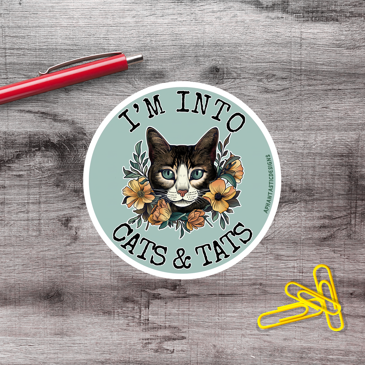A sticker featuring a cat's face surrounded by flowers, with the text "I'm into cats & tats" on it. There is a red pen and a pile of yellow paper clips on a wooden surface in the background.