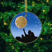 A Christmas ornament with a silhouette of a person pointing upwards, surrounded by a starry night sky and a tree with green needles.