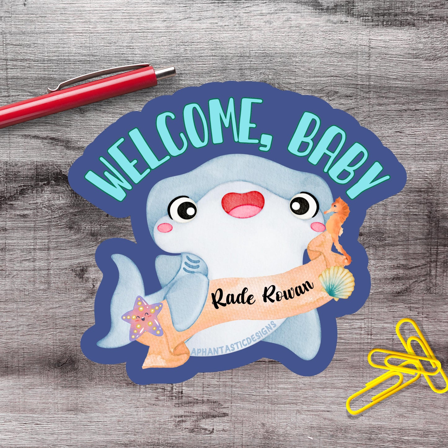 a welcome sign with a cartoon shark and a name tag, placed on a wooden surface alongside a red pen and yellow paper clips.
