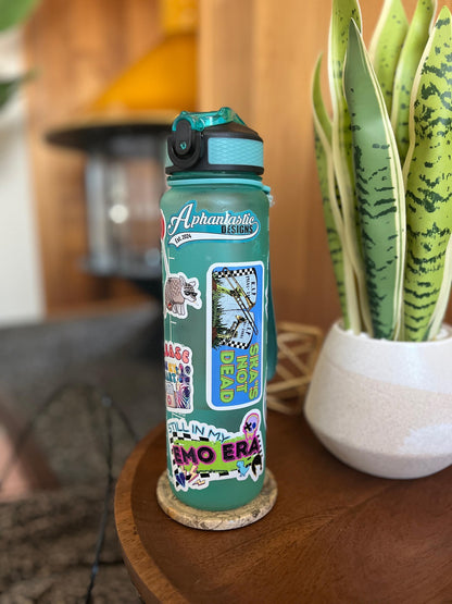 A turquoise water bottle with various stickers and designs on it, placed on a wooden surface next to a potted plant.