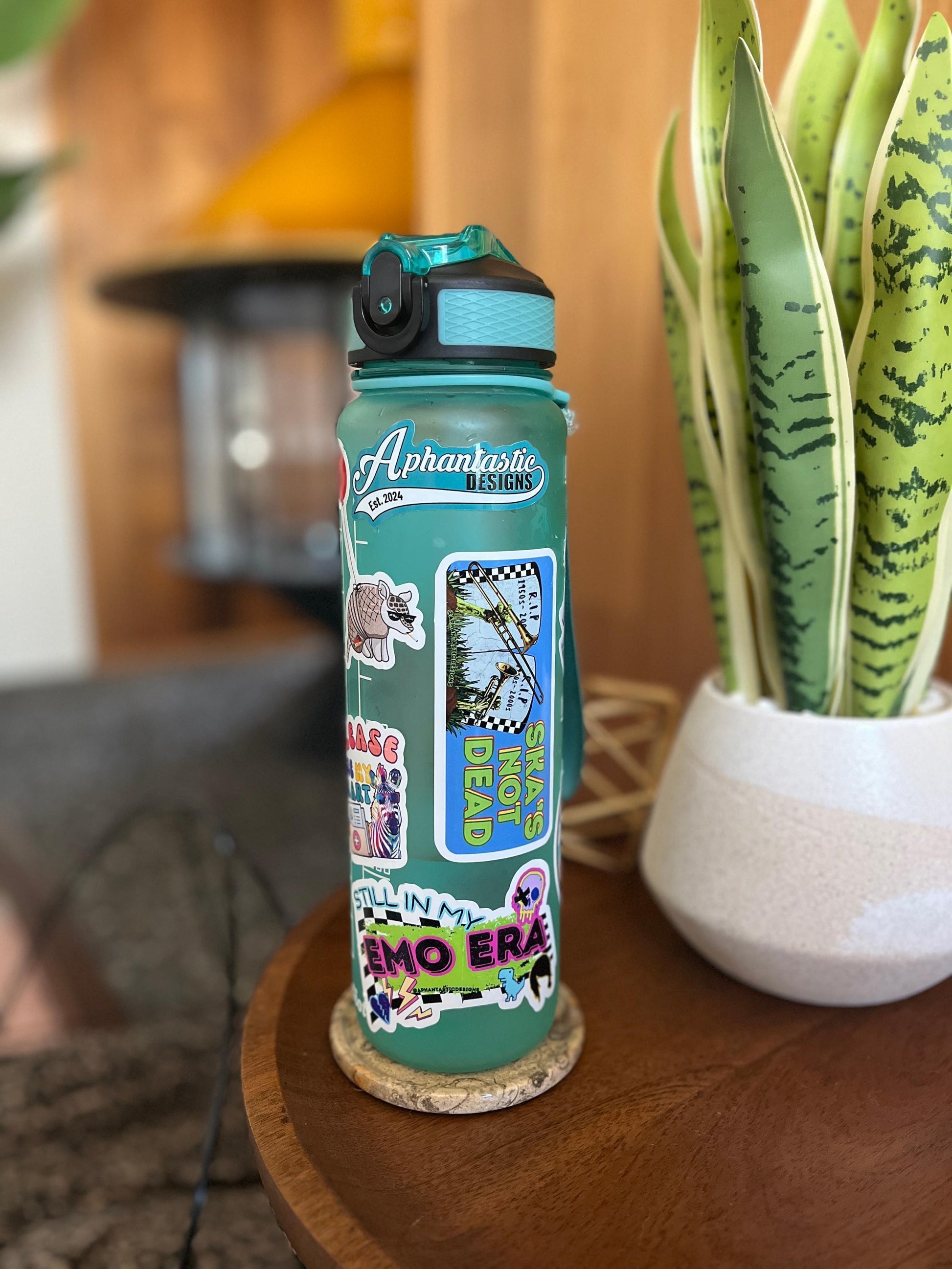 A turquoise water bottle with various stickers and designs on it, placed on a wooden surface next to a potted plant.