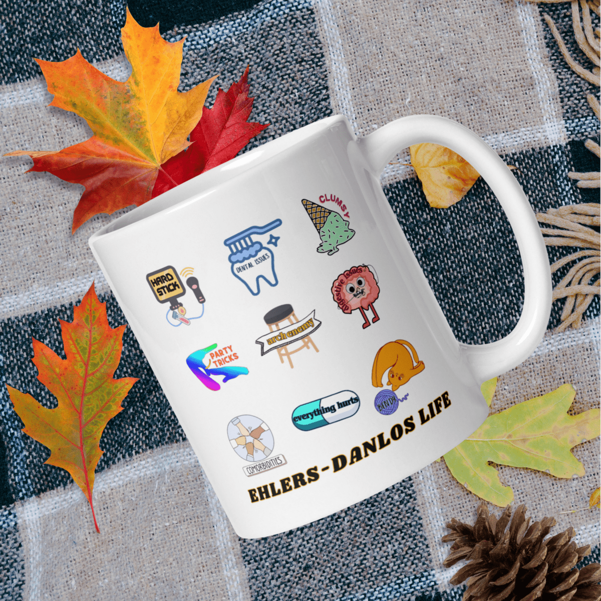 A white mug with various colorful stickers and illustrations on it, placed on a table with autumn leaves and pine cones.