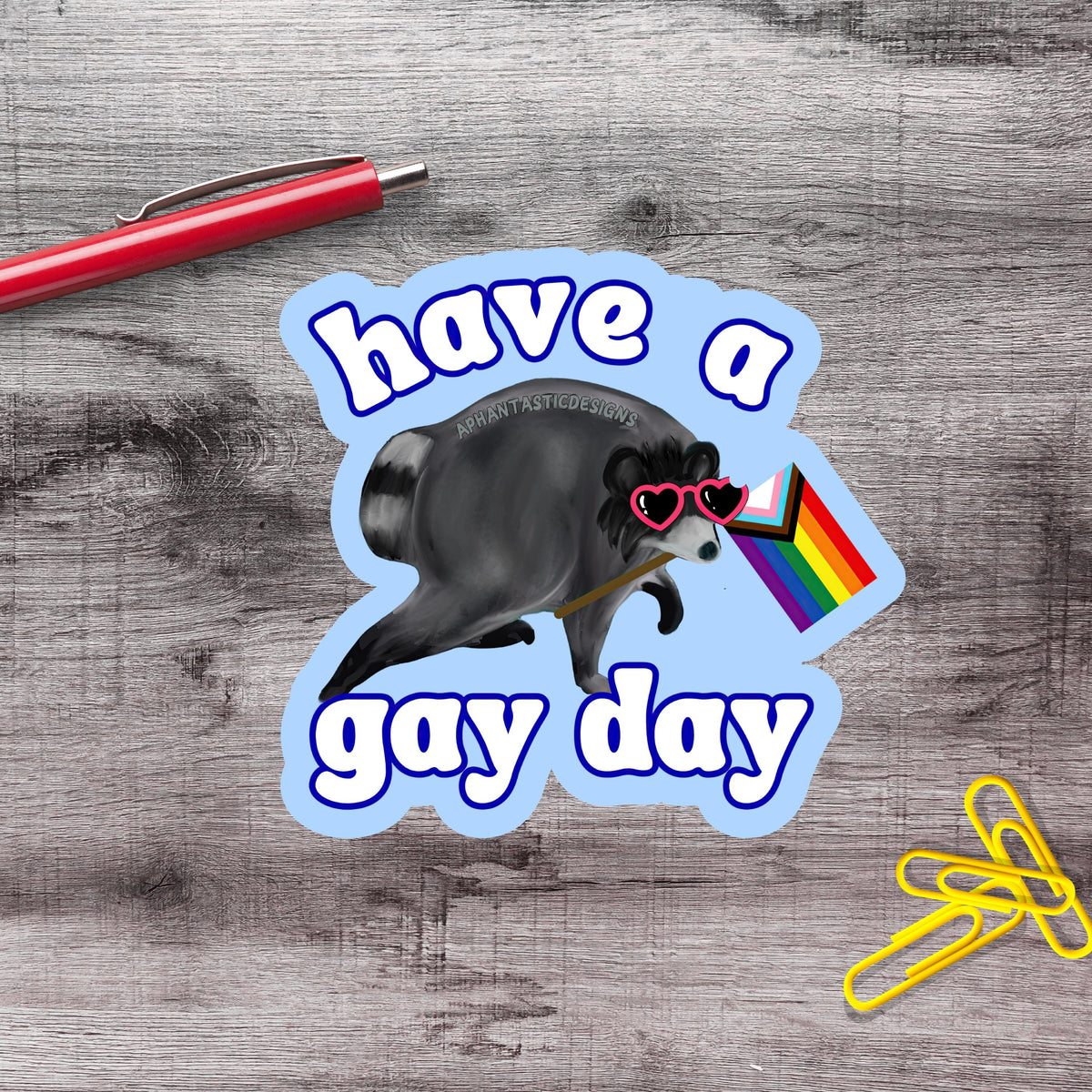 A sticker with a raccoon wearing sunglasses and holding a rainbow flag, along with the text "Have a gay day".