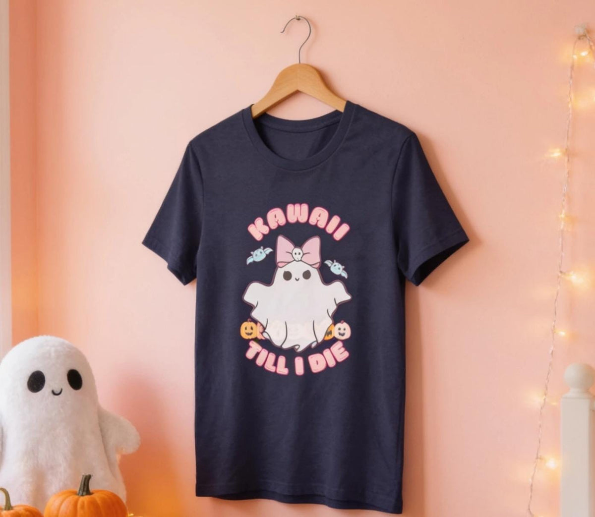 A black t-shirt with a graphic design featuring a ghost character and the text "KAWAII TILL I DIE" is hanging on a wooden hanger against a pink wall.