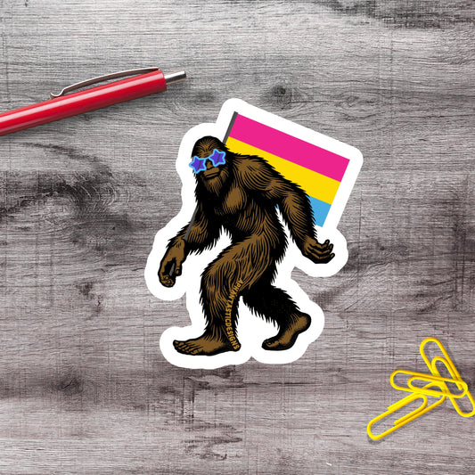 a sticker of a cartoon Sasquatch wearing sunglasses and holding a rainbow flag, along with a red pen and yellow paper clips on a wooden surface.