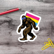 a sticker of a cartoon Sasquatch wearing sunglasses and holding a rainbow flag, along with a red pen and yellow paper clips on a wooden surface.