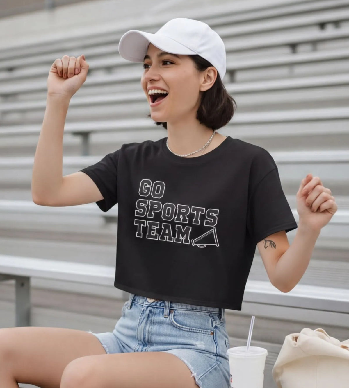 A young woman sitting on bleachers, wearing a black t-shirt with the text "GO SPORTS TEAM" and denim shorts, with a white cap on her head. She is smiling and has her fist raised in the air.