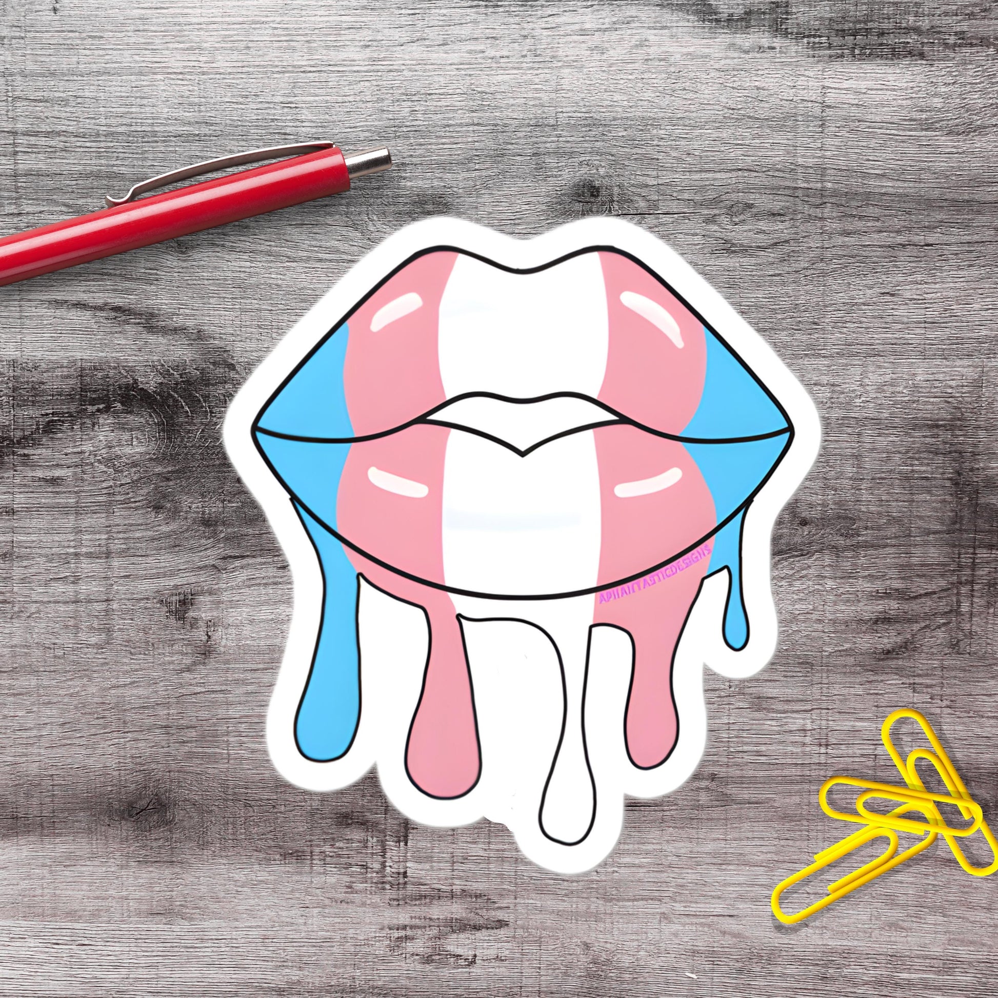 a sticker of a stylized pair of lips with dripping colors, accompanied by a red pen and yellow paper clips on a wooden surface.