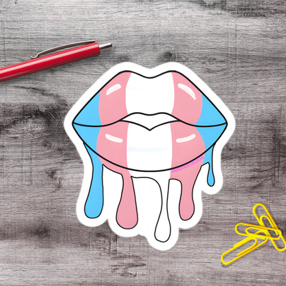 a sticker of a stylized pair of lips with dripping colors, accompanied by a red pen and yellow paper clips on a wooden surface.