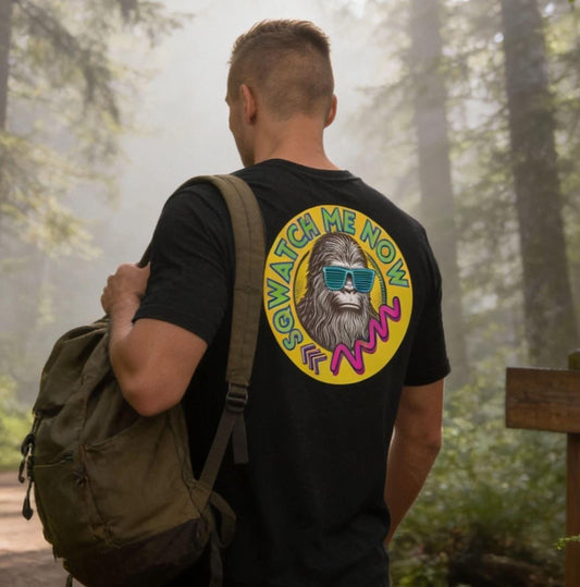 A person wearing a black t-shirt with a colorful graphic and a backpack is walking on a dirt path through a lush, green forest.