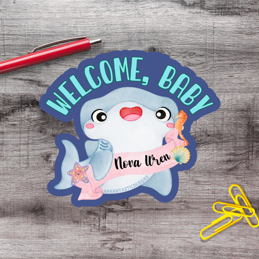 a welcome sign with a cartoon shark and a name tag, placed on a wooden surface alongside a red pen and yellow paper clips.
