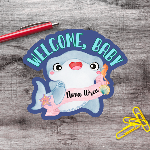 a welcome sign with a cartoon shark and a name tag, placed on a wooden surface alongside a red pen and yellow paper clips.
