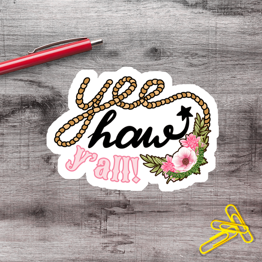 A sticker with the text "Yeehaw Y'all!" written in a decorative font, surrounded by a floral design. A red pen and yellow paper clips are also present on a wooden surface.