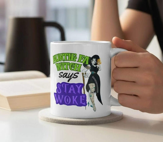 A person is holding a white mug with a colorful design featuring a witch and the text "Auntie Fat Witch says 'Stay Woke'".