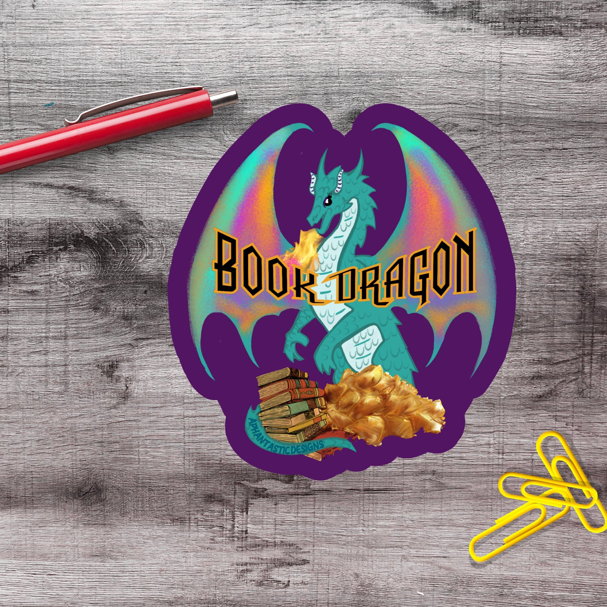 a sticker with a dragon and a pile of books, along with a red pen and yellow paper clips on a wooden surface.