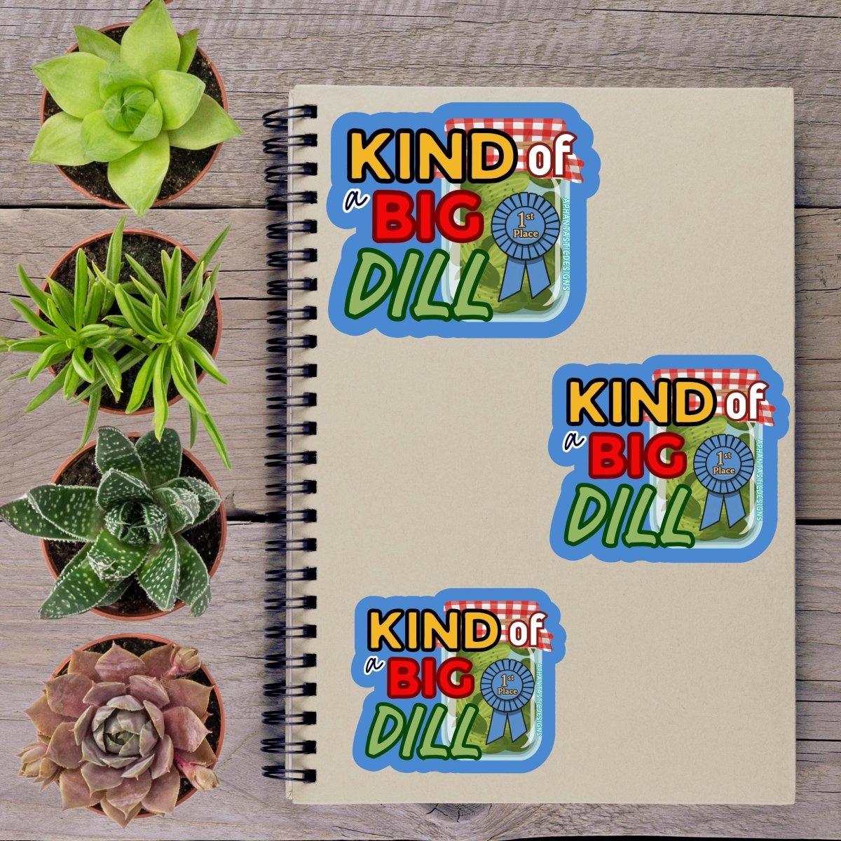 a spiral notebook with stickers on it, surrounded by four potted succulent plants.