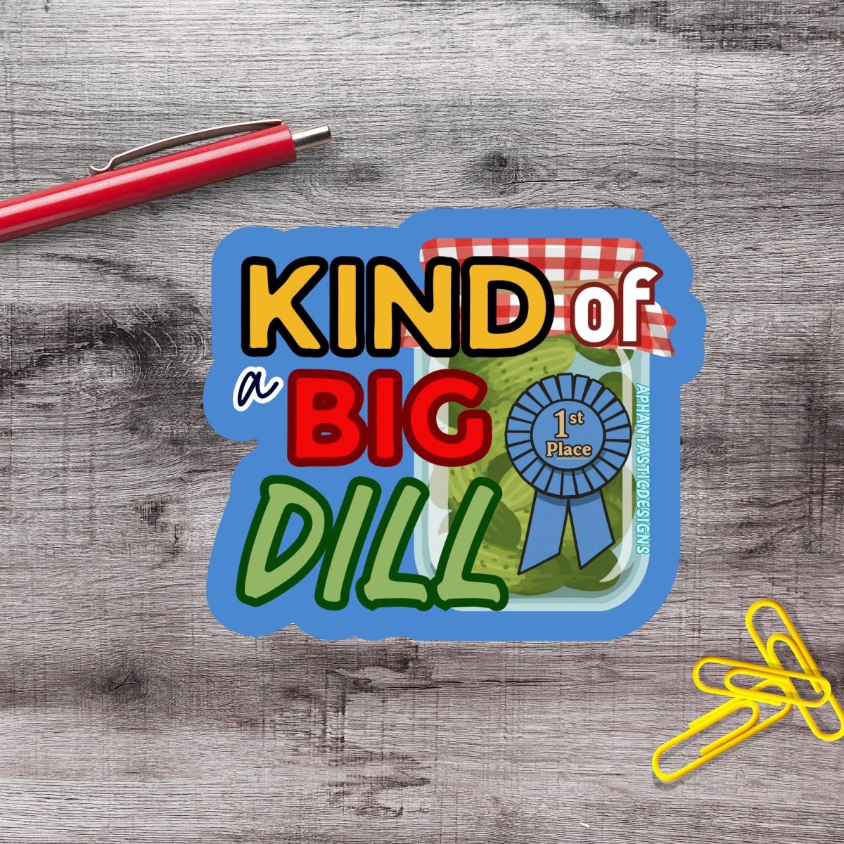 a sticker with the text "KIND of a BIG DILL" and a ribbon, placed on a wooden surface alongside a red pen and yellow paper clips.
