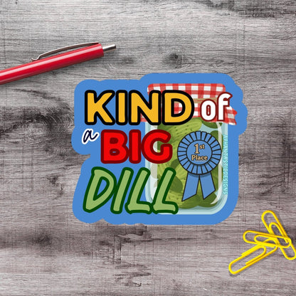 a sticker with the text "KIND of a BIG DILL" and a ribbon, placed on a wooden surface alongside a red pen and yellow paper clips.