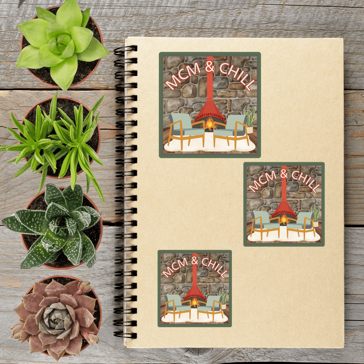 a spiral-bound notebook with three stickers on it, each depicting a cozy scene with a fireplace and chairs. The stickers are arranged on a wooden surface, surrounded by several potted plants.