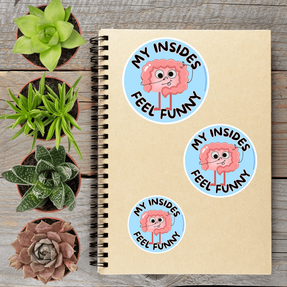 a spiral notebook with stickers on it, surrounded by four potted plants.