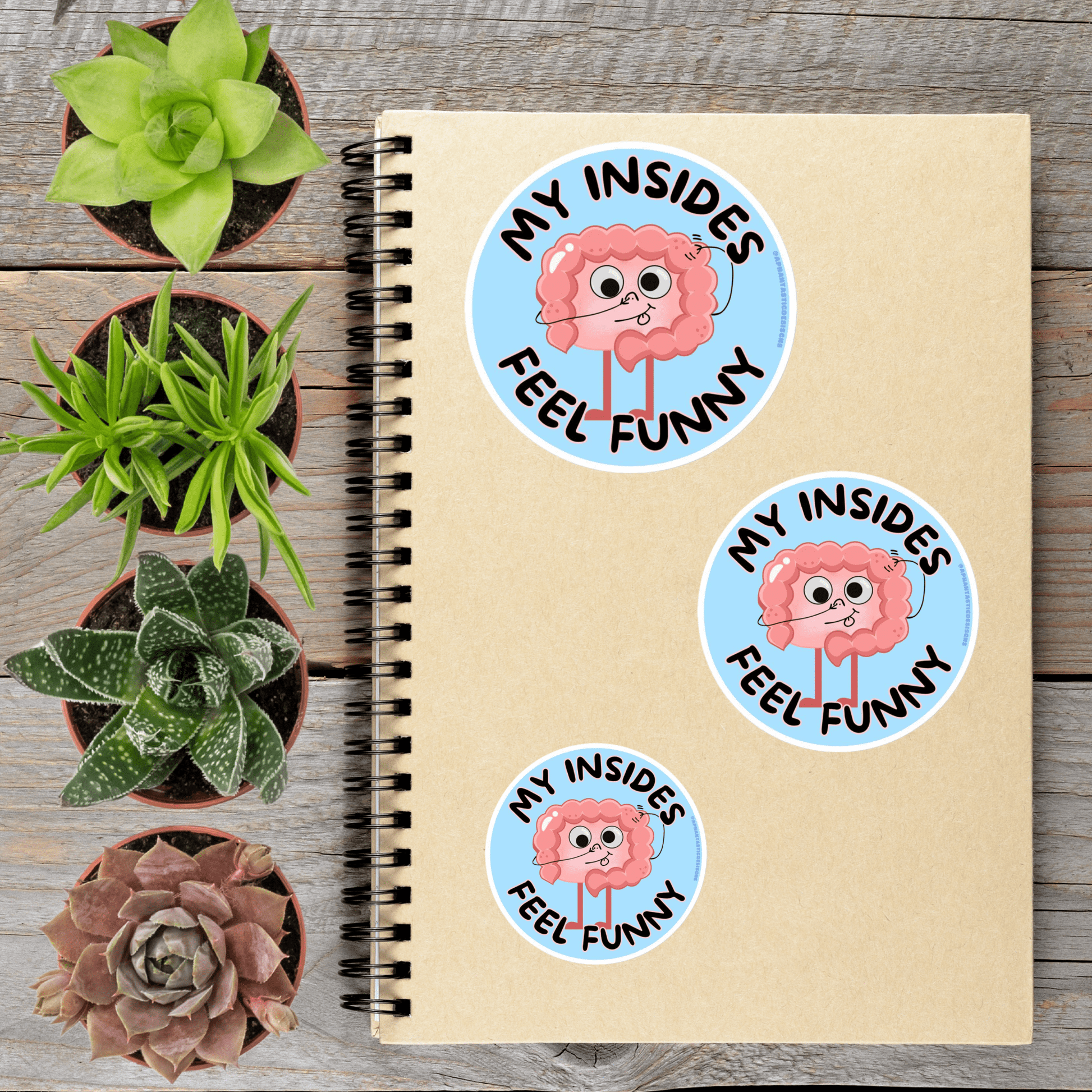 a spiral notebook with stickers on it, surrounded by four potted plants.