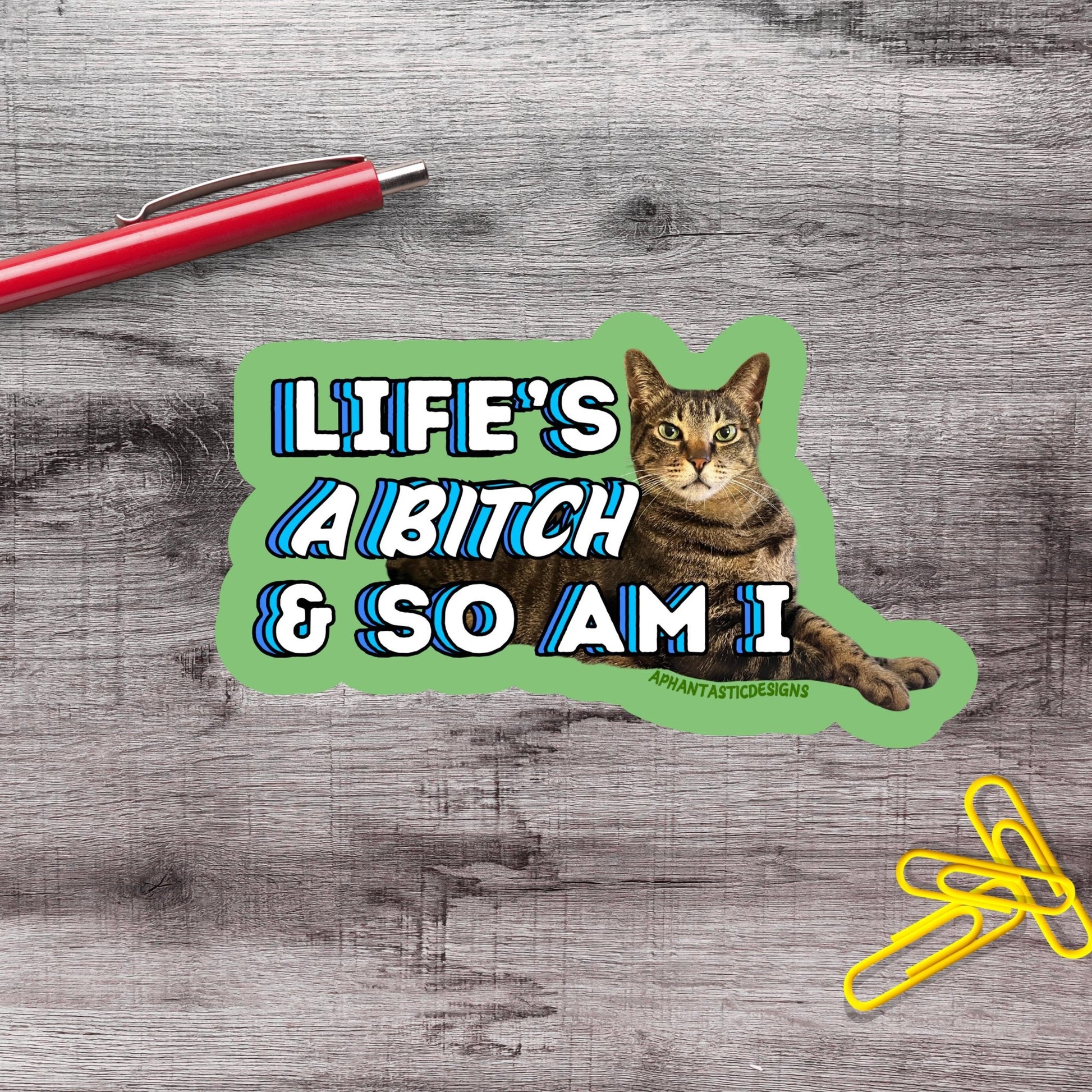 A sticker with a cat and the text "LIFE'S A BITCH & SO AM I" on a wooden surface.