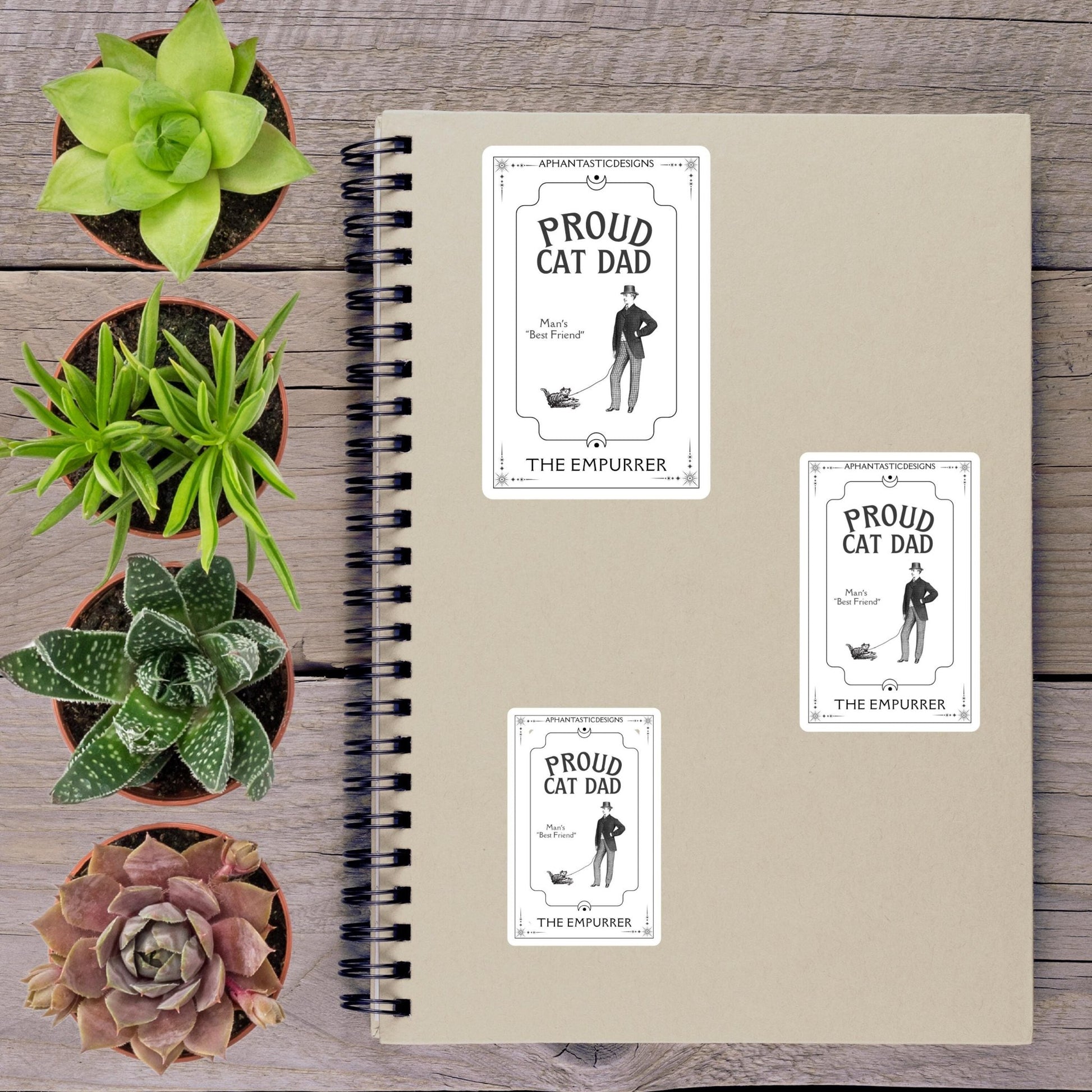 a spiral notebook with three stickers on it, placed on a wooden surface alongside several potted plants.