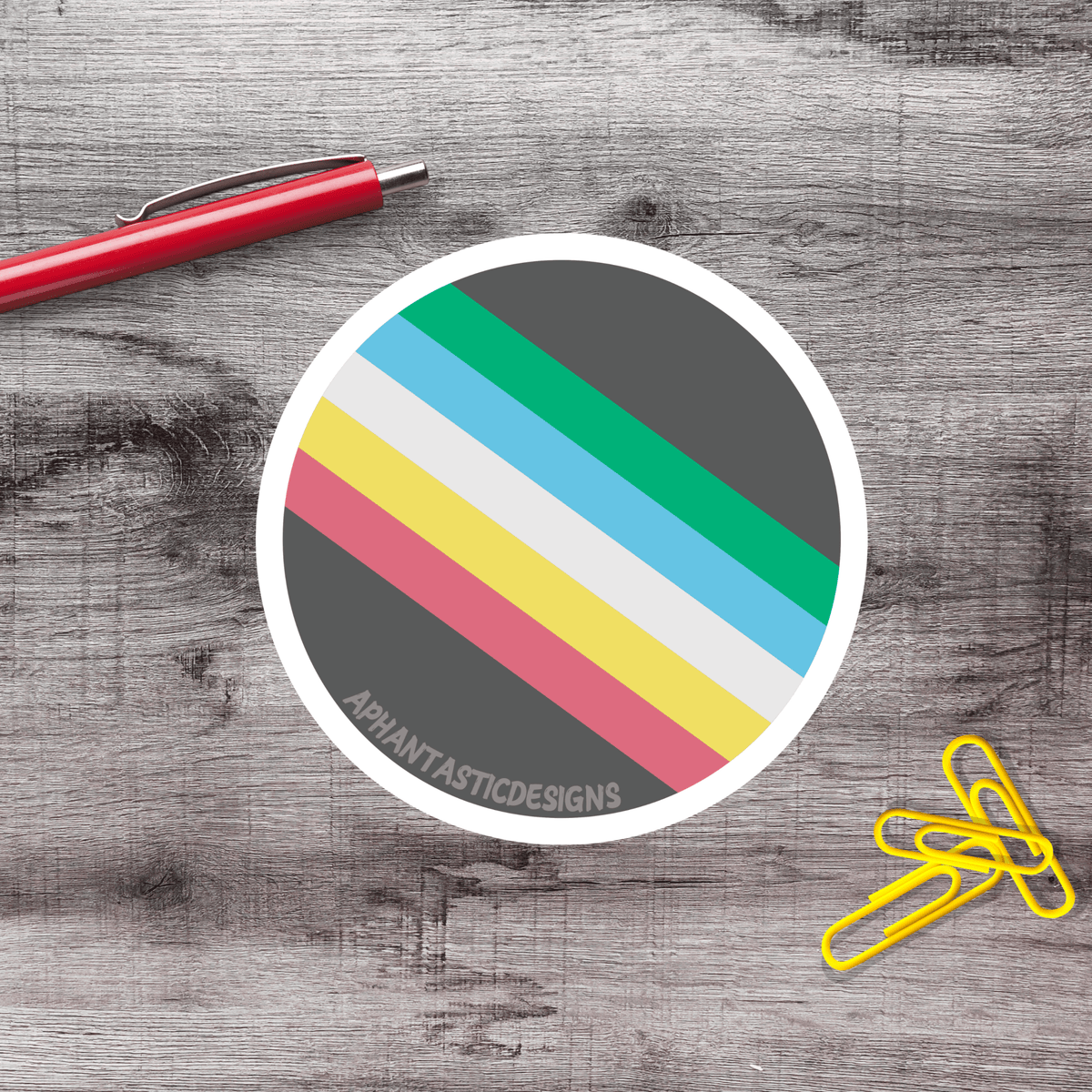 a circular sticker with a colorful striped pattern, a red pen, a stack of yellow paper clips, and a gray wooden surface.