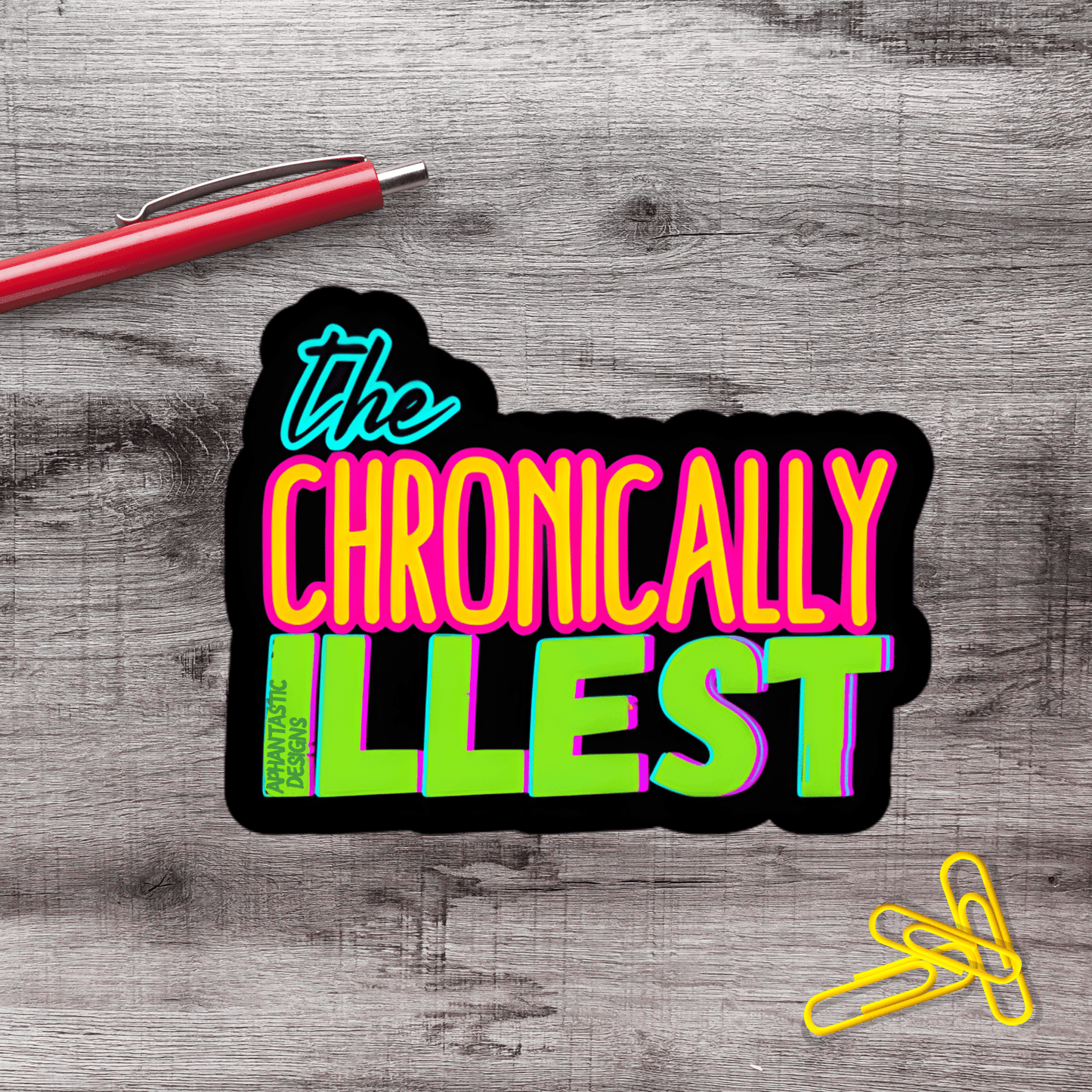 a sticker with the text "The Chronically Ill" in a vibrant, colorful font, placed on a wooden surface alongside a red pen and a pile of yellow paper clips.