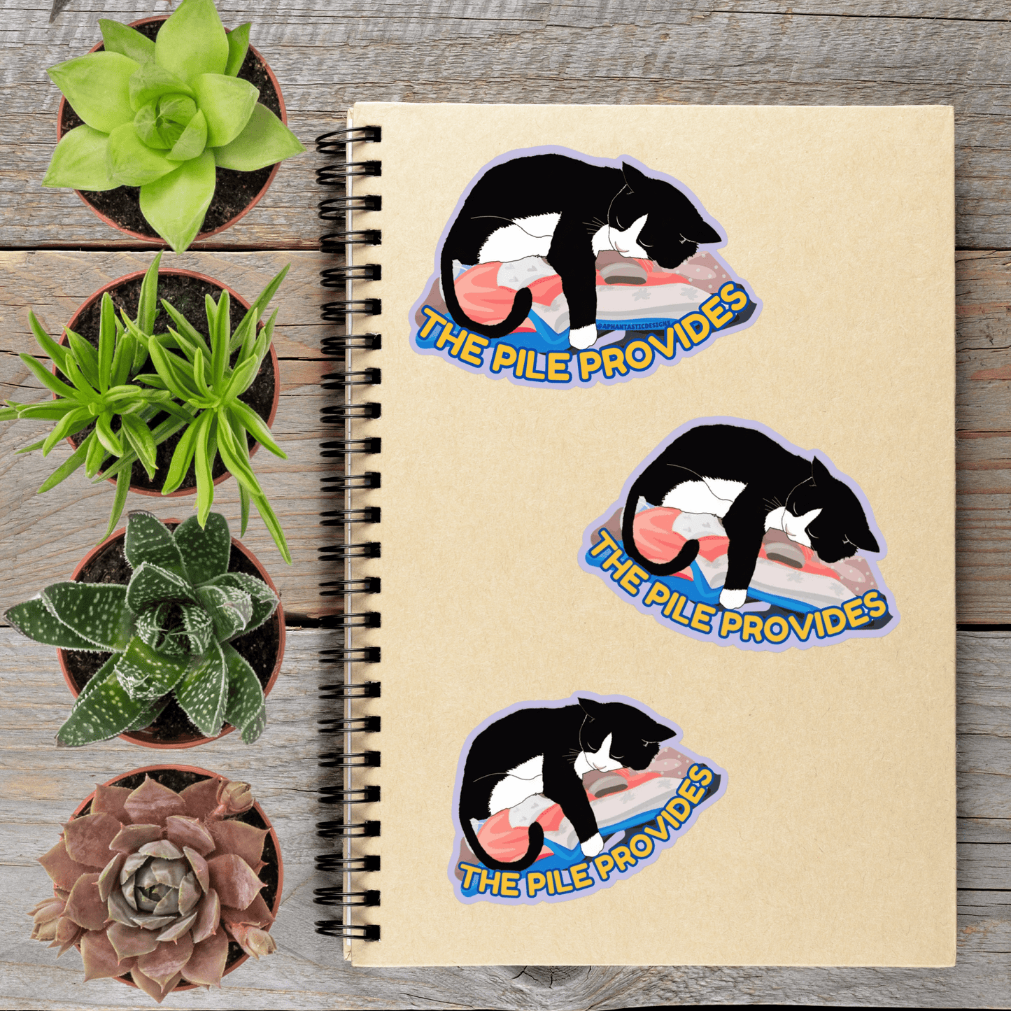 a spiral notebook with three stickers on it, each depicting a black and white cat sleeping on a pile. The stickers are arranged in a row on the notebook, with the first sticker on the left, the second sticker in the middle, and the third sticker on the right. The stickers are placed next to several potted plants, creating a visually appealing and organized display.